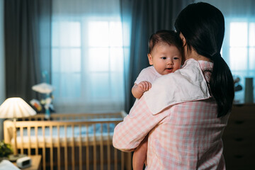 rear view of caring asian mother wearing pajamas with a cloth on shoulder is burping her cute baby daughter in a dark bedroom at night.