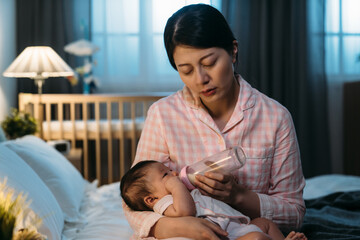 exhausted asian mother is sleeping dozing off while sitting at bedside giving her young baby bottled milk late at night in a dark bedroom with dim light at home.