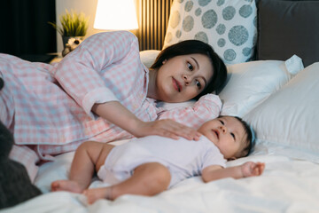 selective focus asian first time mom lying on bed in pajamas is looking at new baby lovingly while putting it to sleep by patting on its chest during nighttime