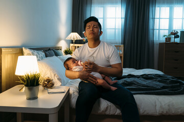 tired asian father sitting at bedside with eyes closed is humming lullaby while holding and feeding...