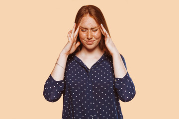 Upset stressed young redhead woman with freckle massaging temples feeling terrible headache migraine, sad tired sick caucasian girl isolated on beige studio background.