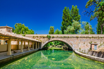 The ancient wash-house and the masonry bridge over the river, in the medieval village of Bevagna. Perugia, Umbria. Blue sky in a sunny summer day. Green algae on the surface of the stagnant water. - obrazy, fototapety, plakaty