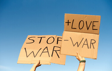 Two Cardboard Signs saying more love, less war, and Stop War. Held by hands on sky background