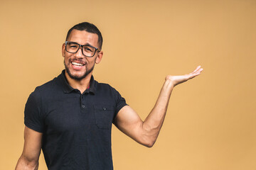 Portrait of happy young african american black man with in casual smiling, pointing aside with finger, looking in camera with excited face expression isolated over beige background.