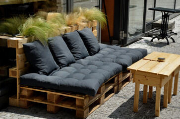 a bench, a sofa, on the street, made from recycled pallets and a few pillows, in front of a store...