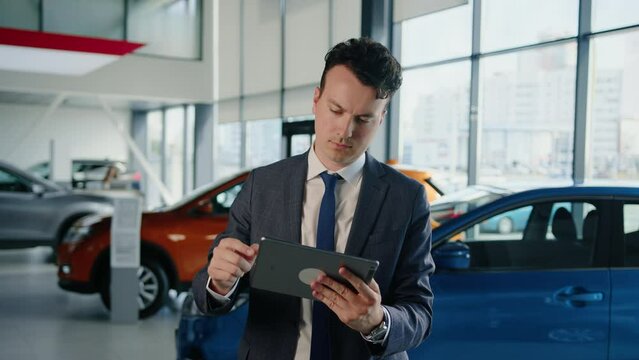 The general manager in a suit and tie with a tablet in his hands on the background of a car dealership