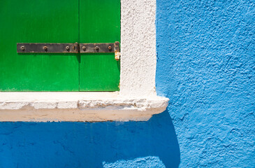 Part of a window with a white border and bright green wooden shutters against a bright blue painted wall