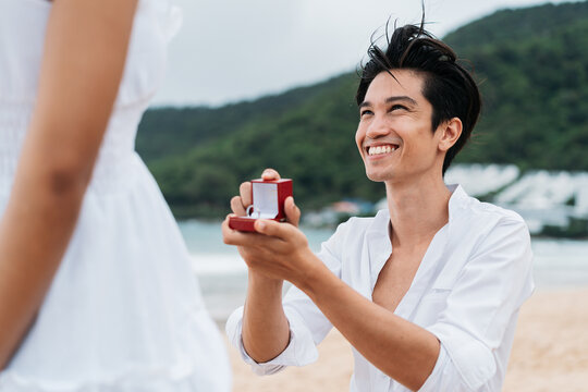 Happy Smiling Asian 30s Man Proposing Woman With A Wedding Ring On The Beach In Summer. A Couple Wearing A White Shirt And Dress - Relationship Goals And Marriage Proposal Concept