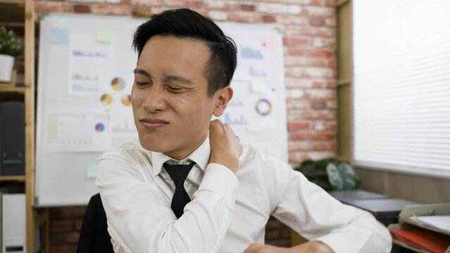 closeup tired Korean male in business wear is exercising his shoulders and neck to relieve stiffness with a painful face at a creative loft office background.