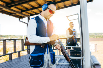 Two adult mans in sunglasses, protective headphones and a rifle vest practicing fire weapon shooting.  - obrazy, fototapety, plakaty