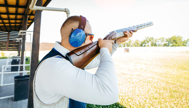 Adult man in sunglasses, protective headphones and a rifle vest practicing fire weapon shooting. Young experienced male aiming shotgun in outdoor.