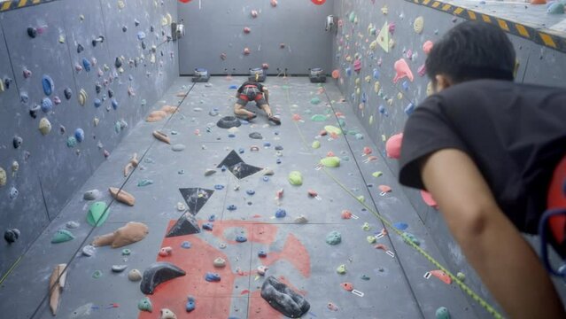 Rock climber on indoor wall belayed by friend at bottom; leisure activity