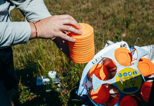 Close up hands of worker loading plat machine with orange shooting plate for shooting-ground training on field with grass