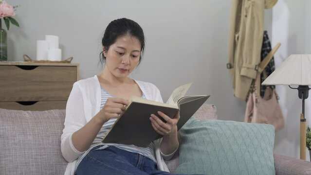 portrait elegant asian young lady is turning pages while reading a hardcover book alone on the sofa in the living room on a tranquil morning.