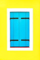 One window with a white border is covered with wooden blue shutters against a bright yellow painted wall