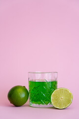 green alcohol drink in faceted glass near fresh limes on pink background.