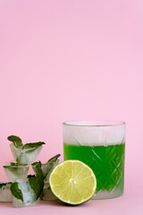 green and fresh lime in faceted glass with alcohol drink near ice cubes with peppermint on pink.