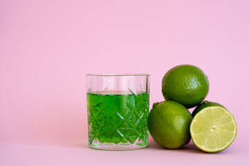 green alcohol drink in glass near fresh and organic limes on pink.