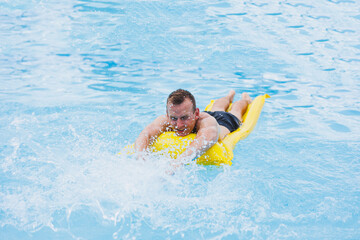 Fototapeta na wymiar A young man in shorts enjoys the water park floating in an inflatable big ring on a sparkling blue pool smiling at the camera. Summer vacation.