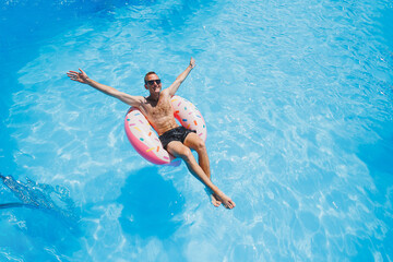 A young man in sunglasses and shorts is relaxing on an inflatable donut in the pool. Summer vacation - obrazy, fototapety, plakaty