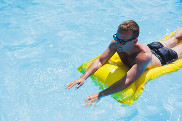 Attractive young man in sunglasses and shorts is relaxing on an inflatable yellow mattress in the...