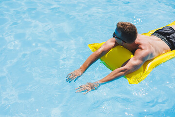 Attractive young man in sunglasses and shorts is relaxing on an inflatable yellow mattress in the pool. Summer vacation