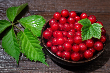 Red currants with leaves in a black bowl on a wooden background. Harvest of ripe summer berries. Closeup
