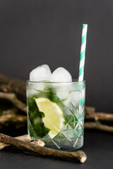 faceted cold glass with ice cubes and sliced lime near wooden sticks on black.