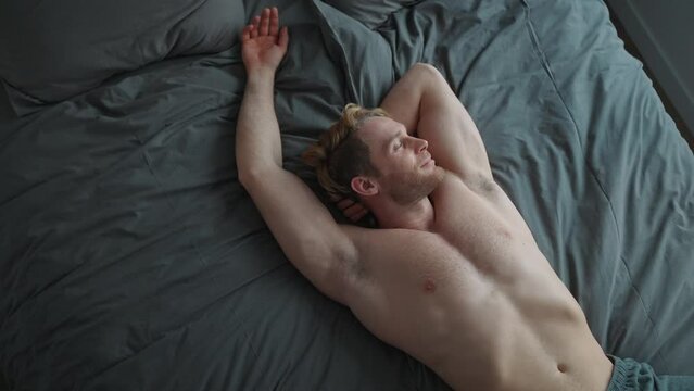 Happy blond athletic man with naked torso lying on the bed at home