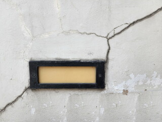 The concrete wall cracks from the top right down to the black square embedded incandescent lamp and diagonally to the floor. The cracks have traces of water seepage. Can see traces of peeling paint.
