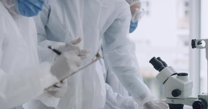 Scientists in hazmat suits using a microscope in a lab. Closeup of a team of microbiologists discussing forensic research while analyzing medical samples and writing their findings to develop a cure