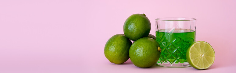 green liquid in glass near fresh limes on pink background, banner.