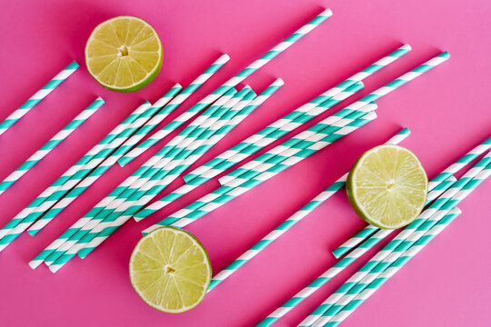 top view of striped blue and white straws near limes on pink background.