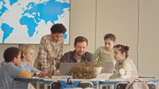 Medium slowmo of diverse school kids and male teacher looking at world map on laptop and having discussion at Geography lesson