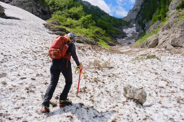 Woman alpinist with ice axe, crampons and backpack, hikes up on late Summer snow in the White Valley, an unmarked alpine route in Bucegi mountains, Romania. - obrazy, fototapety, plakaty