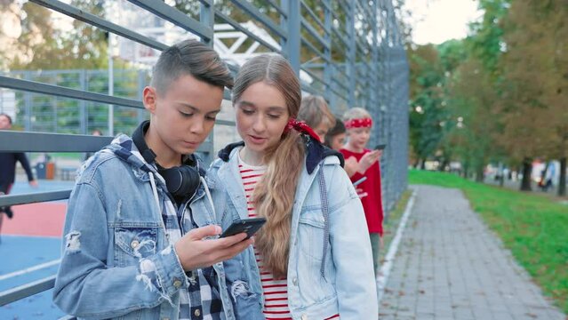 Close up portrait of young cute stylish caucasian teens standing outdoors texting on smartphone and talking looking at screen Boy and girl using mobile phone on street in park, urban lifestyle concept