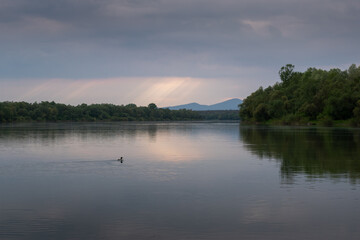Obraz na płótnie Canvas Sun ray piercing through clouds over river, forest on river bank and mountain silhouette in distance