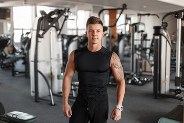 Fototapeta na wymiar Portrait of a handsome fashionable athletic man model with hairstyle and a perfect muscular body in black sportswear with a T-shirt in front of the gym
