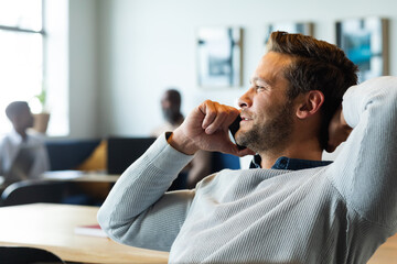 Smiling caucasian mid adult businessman with hand behind head talking on mobile phone in office