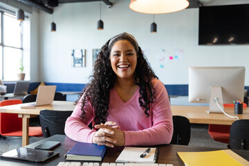 Portrait of happy biracial young businesswoman sitting at desk in creative office