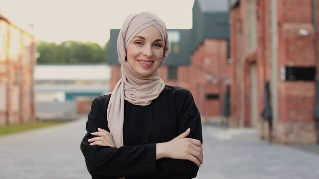 Portrait of young muslim woman wearing hijab head scarf in city while looking at camera. Closeup face of cheerful woman covered with headscarf smiling outdoor. Casual islamic girl in town.