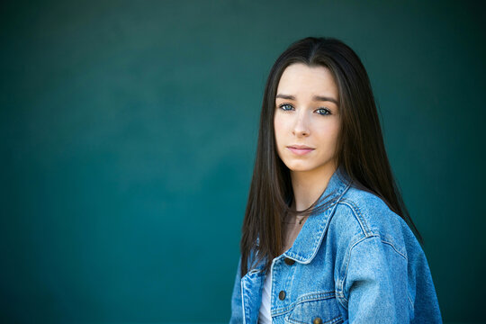 A beautiful teen brunette girl with a serious look leaning against a wall in thought with a teal green turquoise background