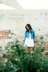 A teenage girl with freckles and long dark stright hair outside leaning against a brick wall and looking away from the camera