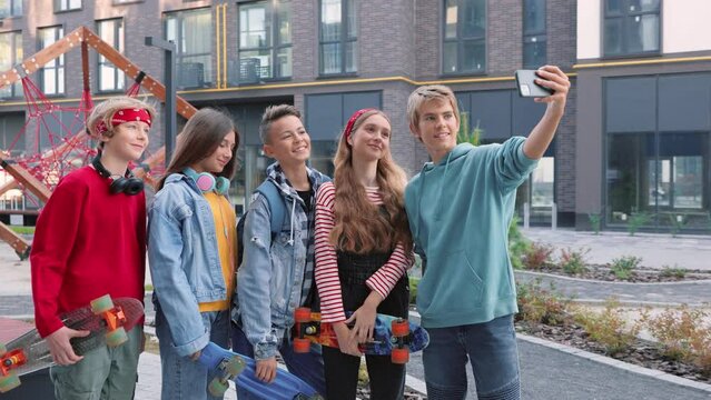 Happy smiling friends stylish teenagers with skateboards and taking selfie photos on smartphone standing outdoor Joyful boys and girls skaters posing to cellphone camera taking pictures, skateboarding