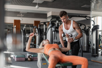 Obraz na płótnie Canvas Athletic woman with a male bodybuilder fitness instructor lying on a bench and doing exercises with dumbbells in the gym. The couple is working out. Guy trains girl