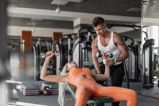 Athletic woman with a male bodybuilder fitness instructor lying on a bench and doing exercises with dumbbells in the gym. The couple is working out. Guy trains girl