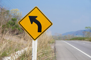 Yellow traffic sign with arrow left curve symbol beside the rural road to warn drivers to know the way ahead has a left curve. Concept : Warning traffic sign for transportation.                      