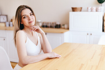 Obraz na płótnie Canvas Young woman is sitting at Wooden Table in front of White Cozy Kitchen. Rent Housing with Good Repair