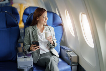 portrait of A successful asian business woman or female entrepreneur in formal suit in a plane sits in a business class seat and uses a smartphone with drink coffee during flight, relax concept