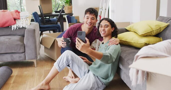 Happy diverse male couple moving house, drinking coffee, taking selfie using smartphone